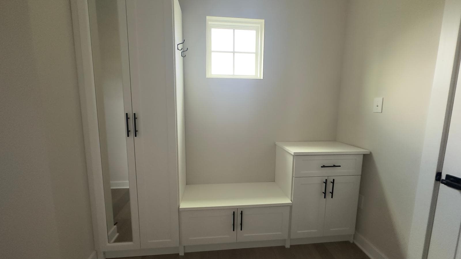 Custom white mudroom entry unit with mirrored wardrobe cabinet, storage bench with coat hooks, and a base cabinet with drawer