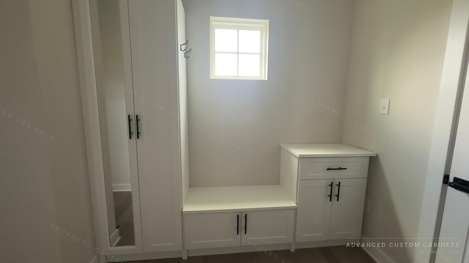 Custom white mudroom entry unit with mirrored wardrobe cabinet, storage bench with coat hooks, and a base cabinet with drawer