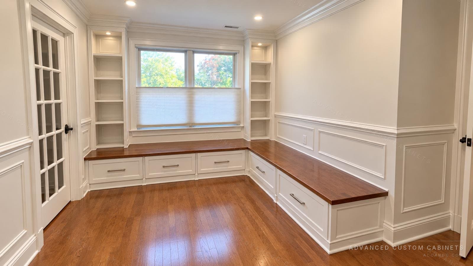 White built-in window seat with a warm wood top over drawers, flanked by built-in bookcases on both sides of the window in a paneled room