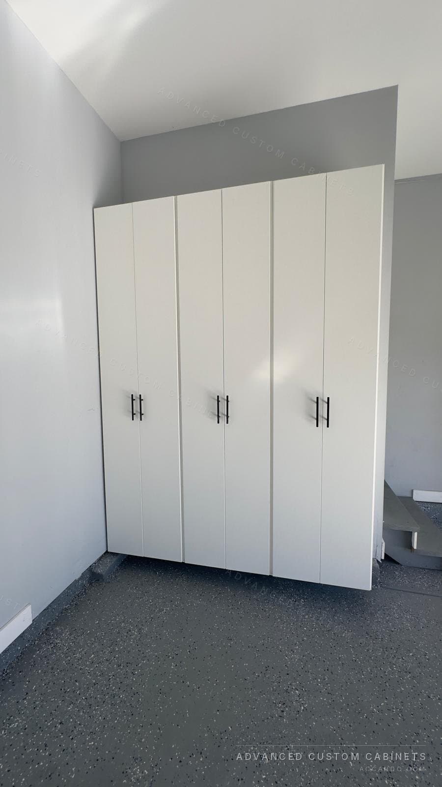 Three tall white double-door garage cabinets with black bar pulls, mounted on a grey wall and raised off a speckled epoxy floor