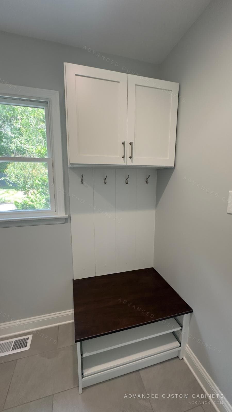 Custom white mudroom locker with an upper cabinet, coat hooks, and a dark-wood bench over open shoe shelves