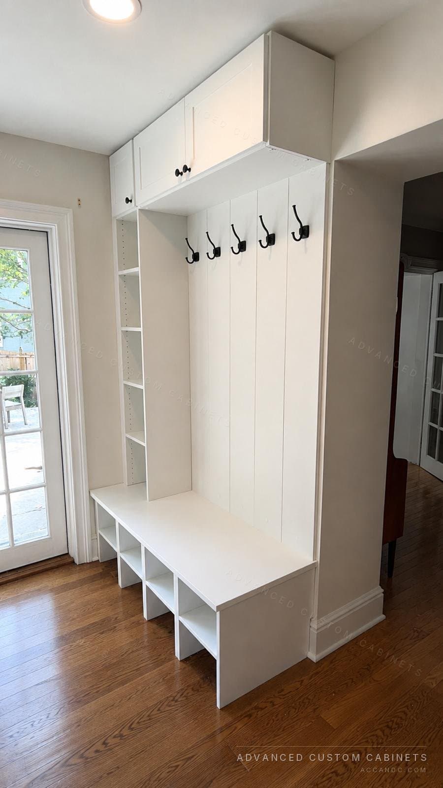 Custom white mudroom bench with shoe cubbies, black coat hooks on shiplap, and an overhead storage cabinet