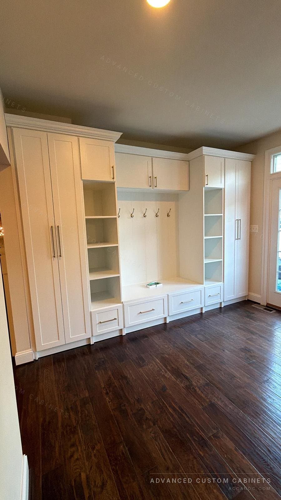 Custom white mudroom built-in with flanking tall wardrobe cabinets, a storage bench with drawers, coat hooks, and open cubbies over dark hardwood