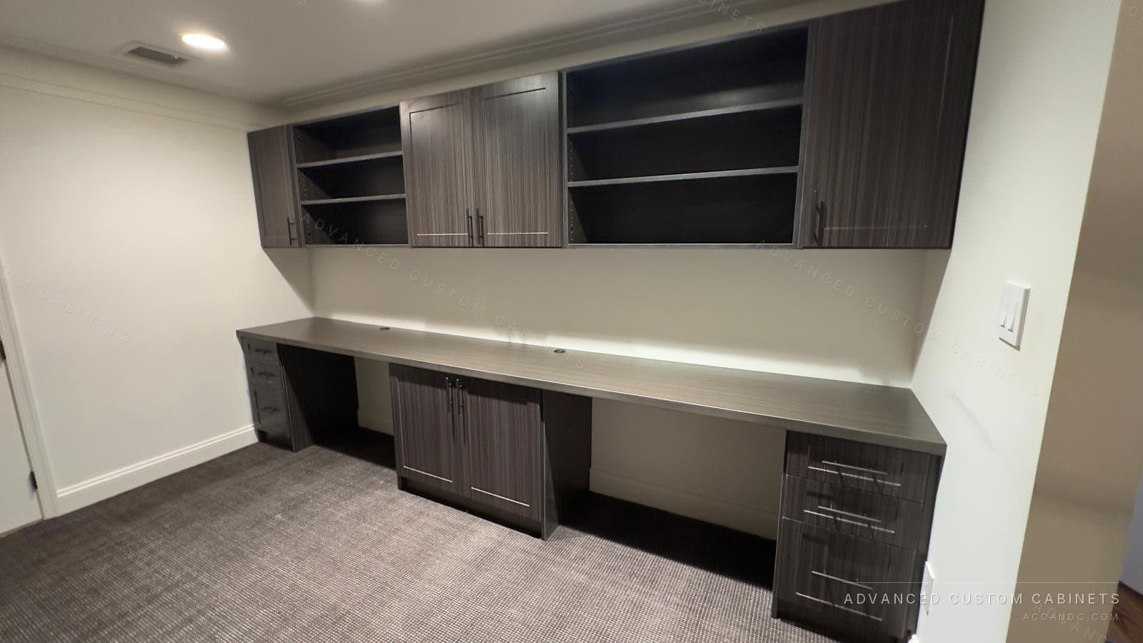 Custom home office wall with a long gray wood-grain desk, drawer pedestals at both ends, and upper cabinets with shaker doors and open shelves
