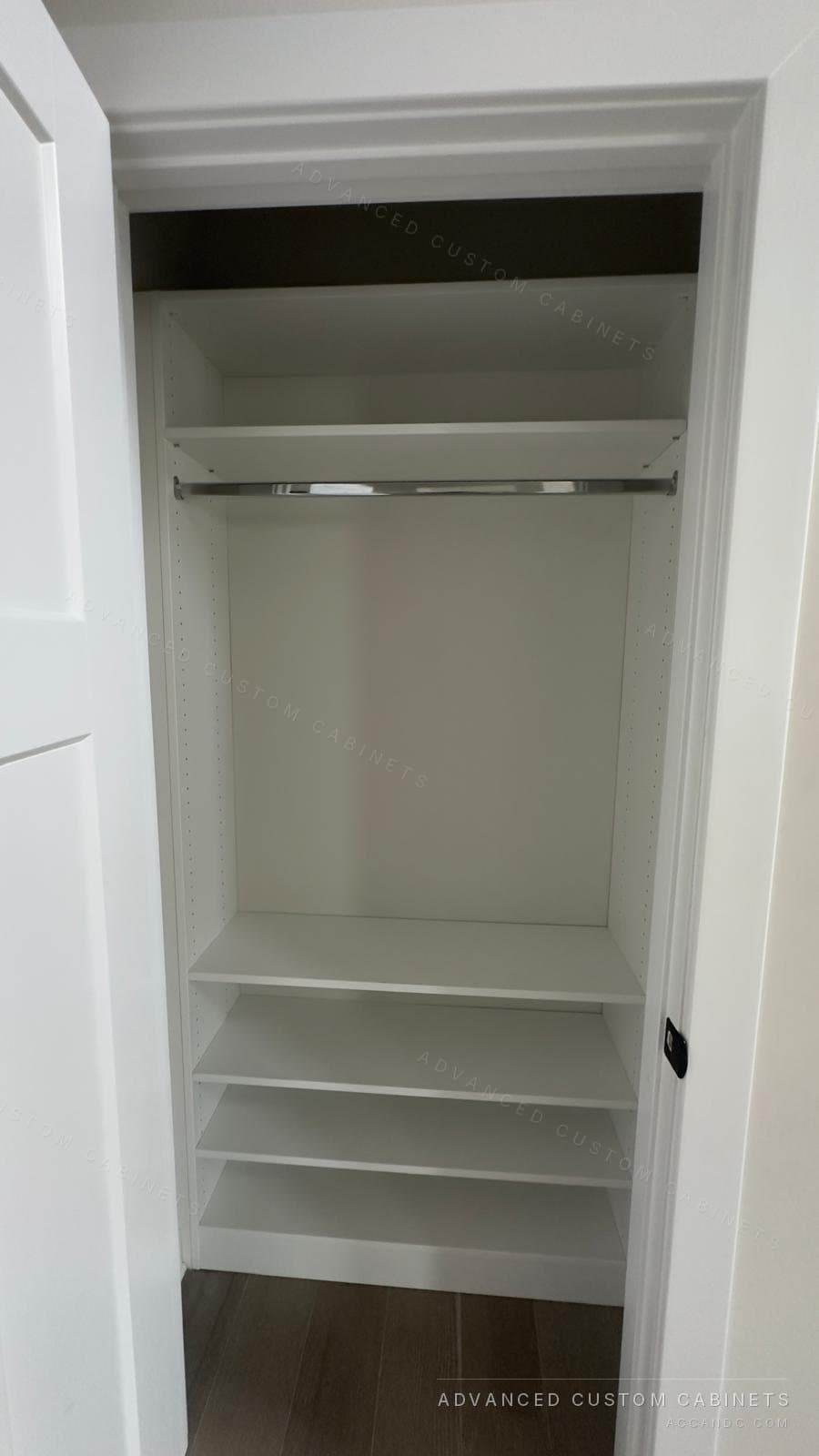 White reach-in closet with a hanging rod, upper shelf and a stack of lower shelves behind a white door frame