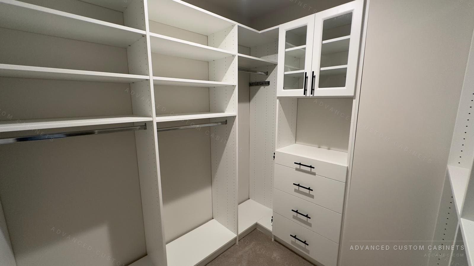 Corner of a white walk-in closet with double hanging under deep shelves and a glass-door cabinet over four drawers with black bar pulls