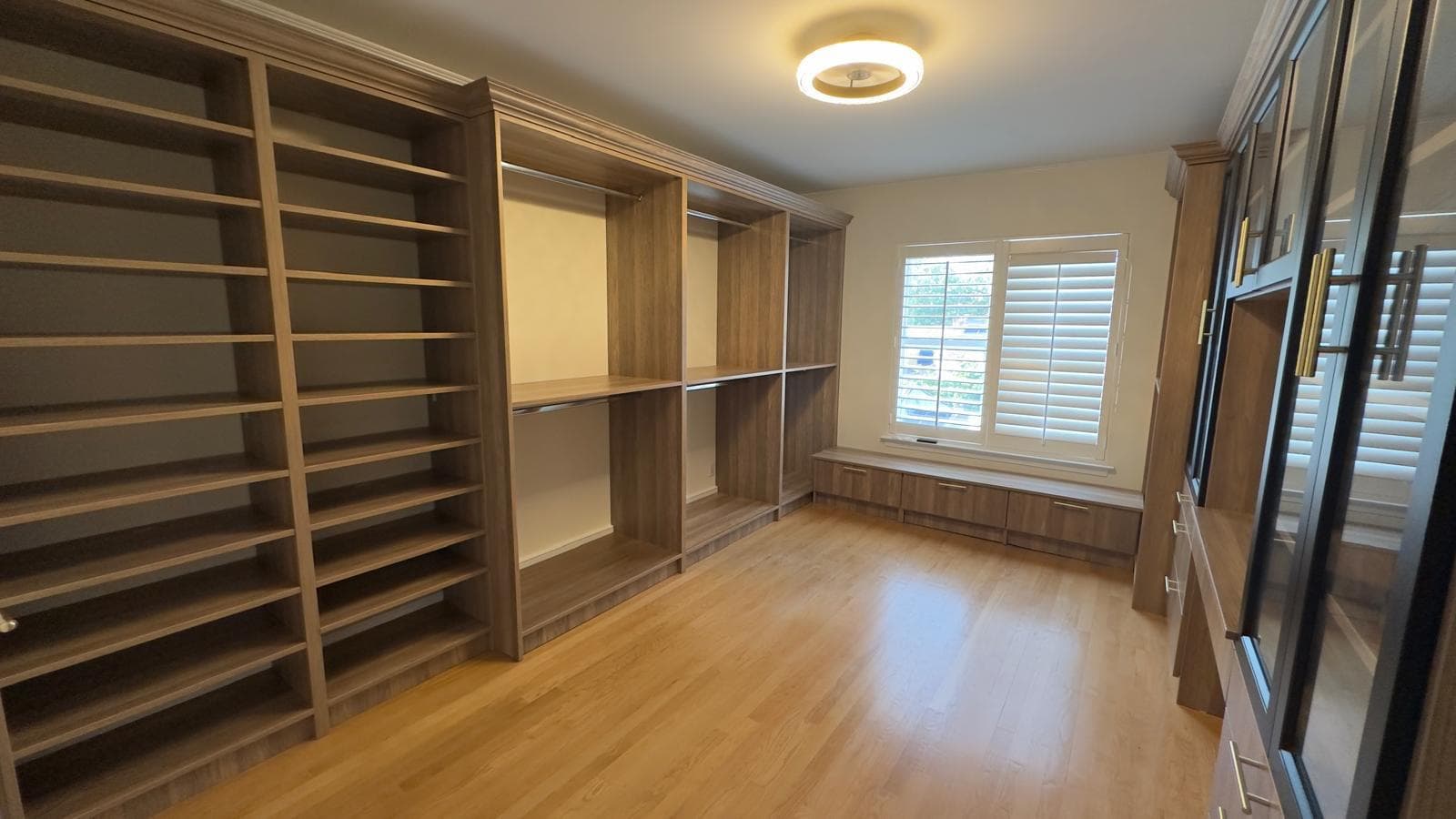 Custom walk-in closet in wood finish with tall open shelving, hanging sections, a window bench, and a black glass-door display cabinet