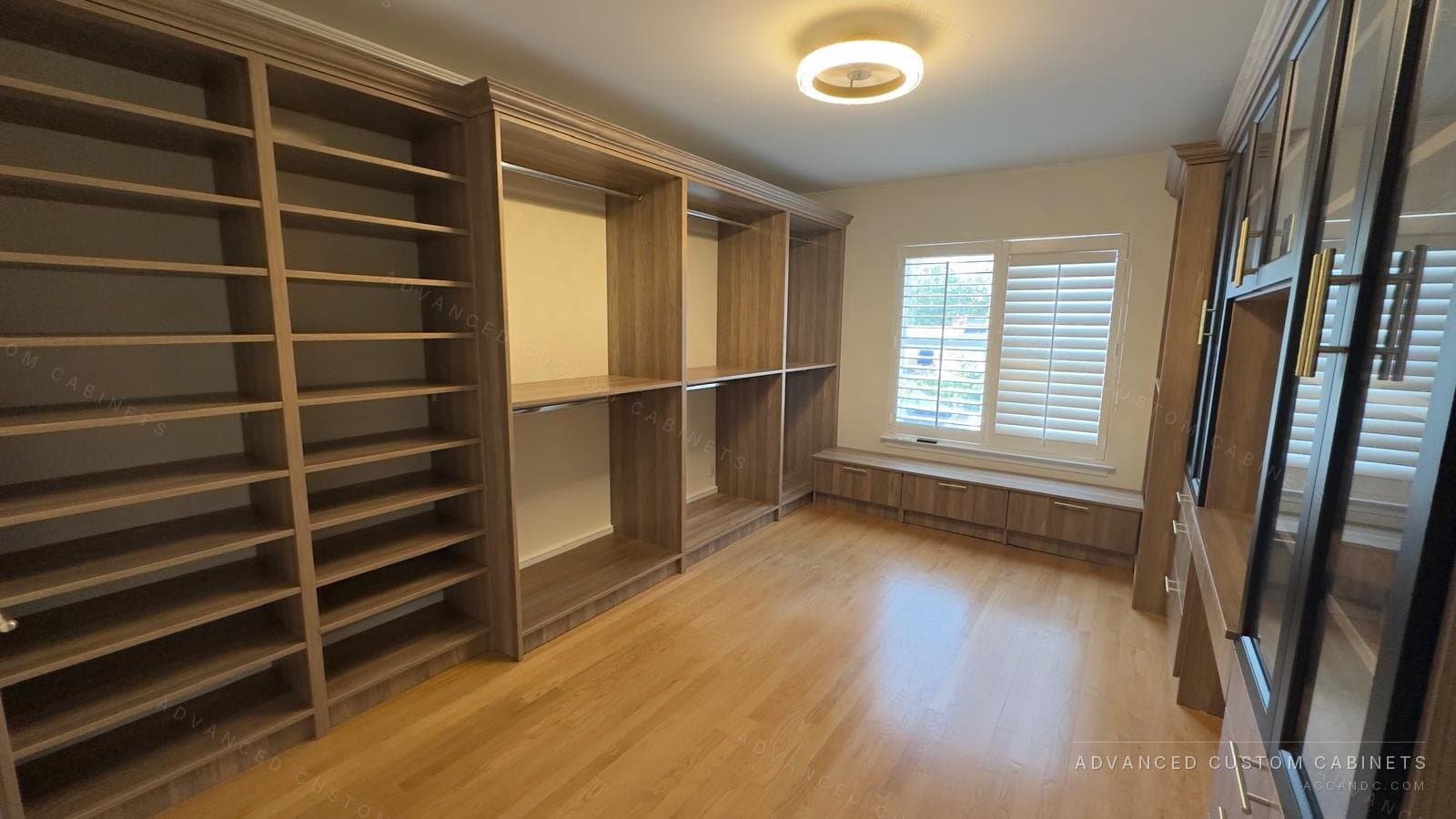 Custom walk-in closet in wood finish with tall open shelving, hanging sections, a window bench, and a black glass-door display cabinet