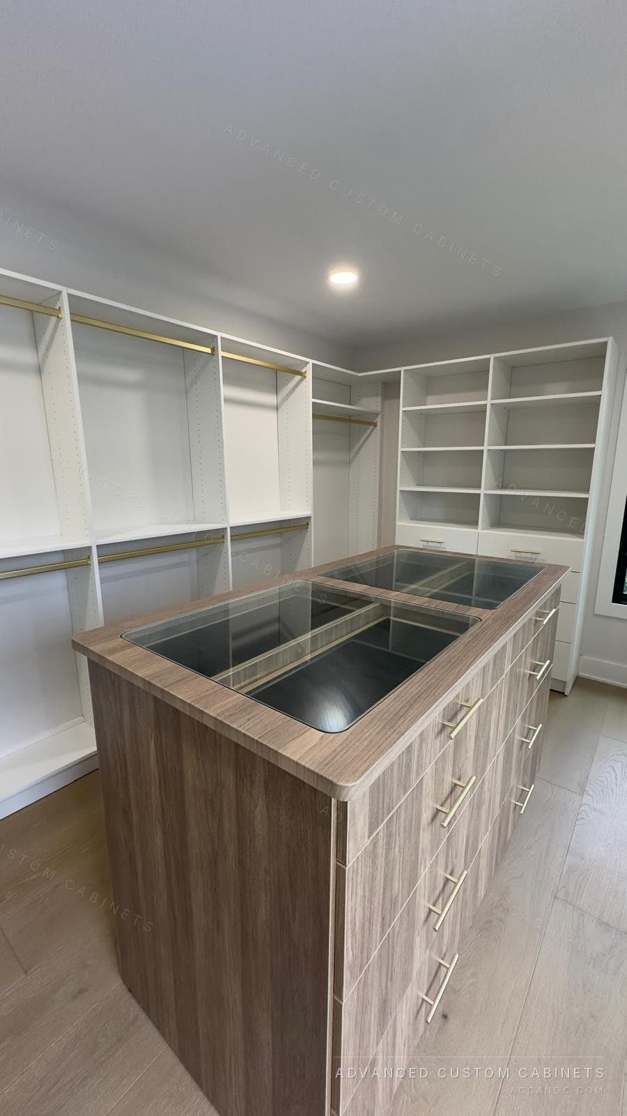 Custom walk-in closet with a wood-front center island under a glass display top, brass hanging rods, and white perimeter shelving