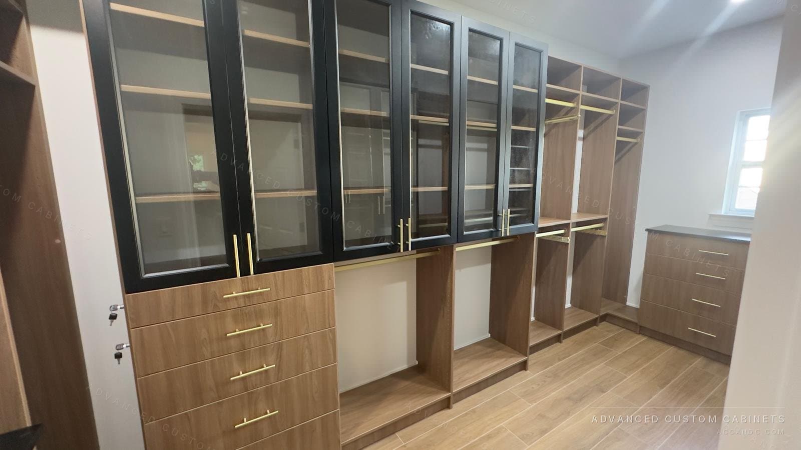 Light oak walk-in closet with black glass-front upper cabinets, brass bar pulls, a drawer bank and open hanging sections under a window