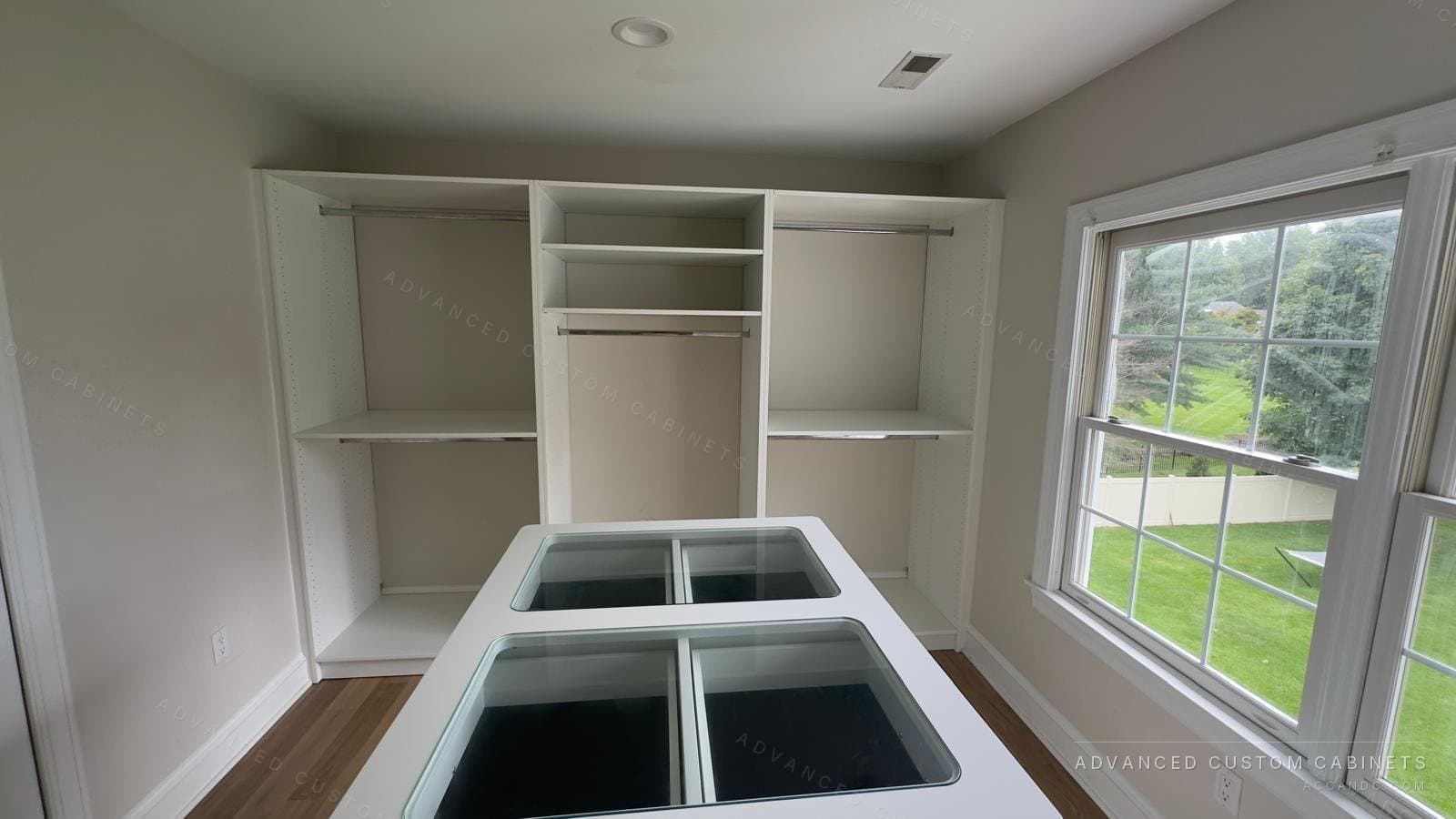 Custom white walk-in closet with a glass-top display island, double hanging sections, and open shelving beside a garden window