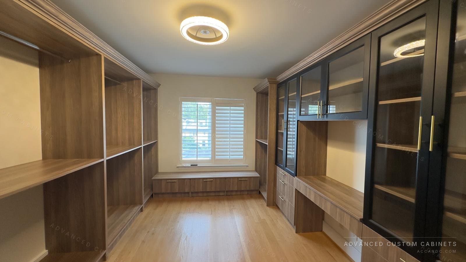Wide view of a custom walk-in closet with wood open shelving on both walls, a window bench, built-in desk, and black glass cabinets