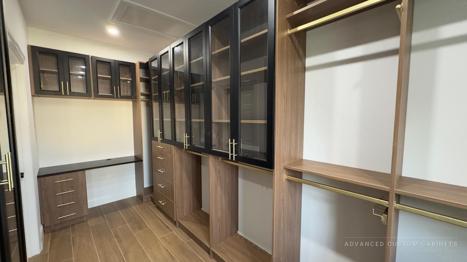 Walk-in closet dressing wall in light oak with glass-front uppers, brass rods, a built-in vanity desk and drawer bases