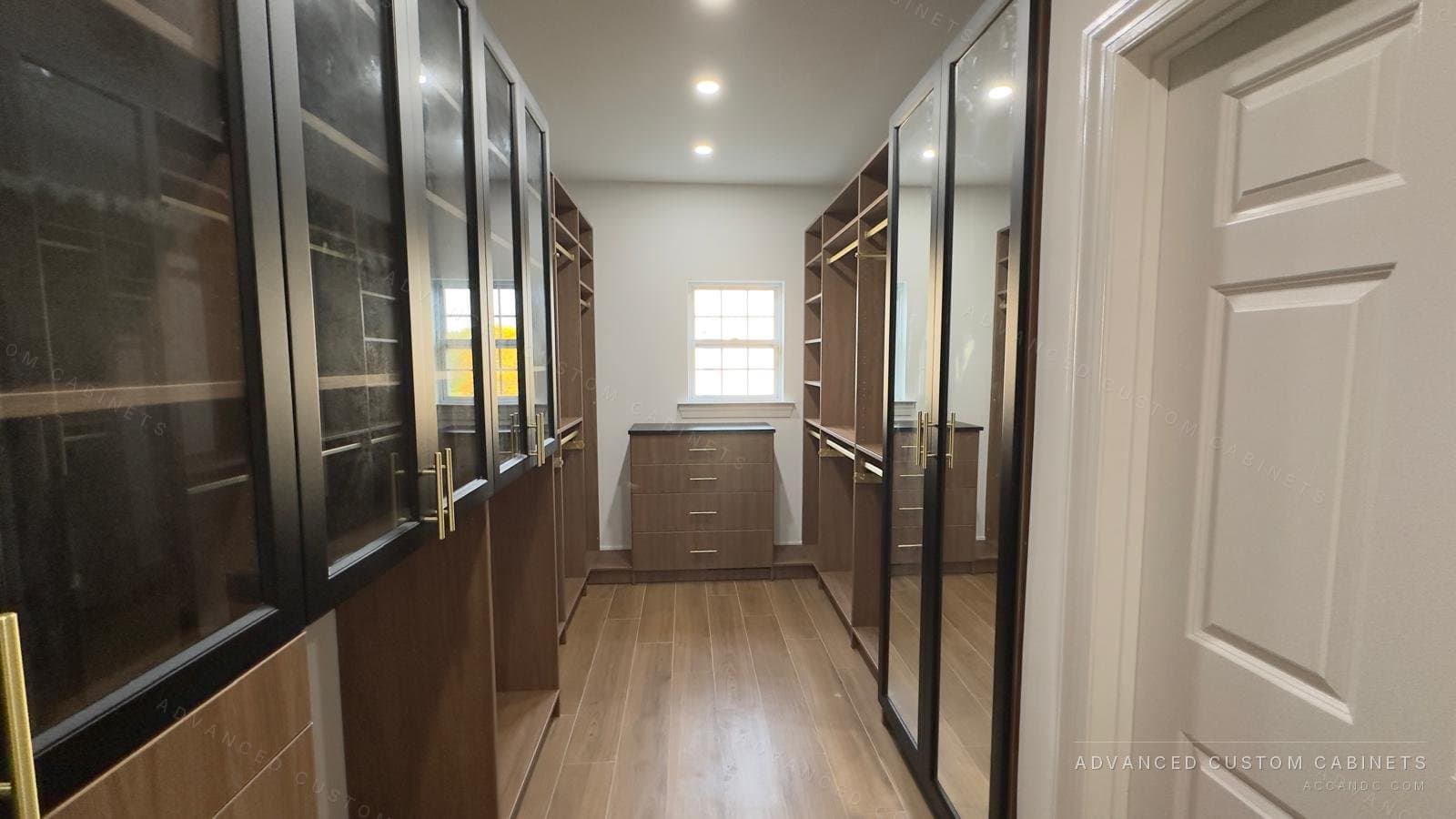 Walk-in closet with black-framed glass-front upper cabinets over light oak drawers, brass pulls and a mirrored sliding door, seen from the hallway