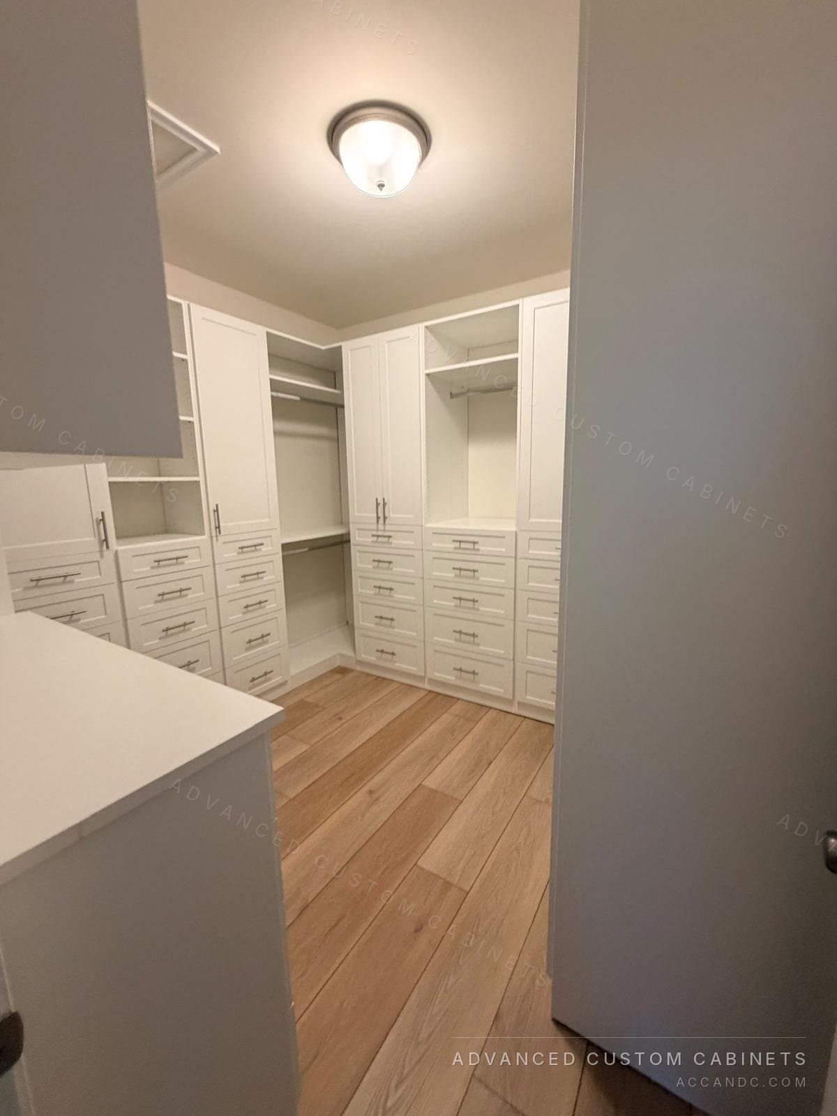 White walk-in closet seen from the doorway, with shaker drawer banks under tall doors and open double hanging around three walls, satin bar pulls and light oak floor