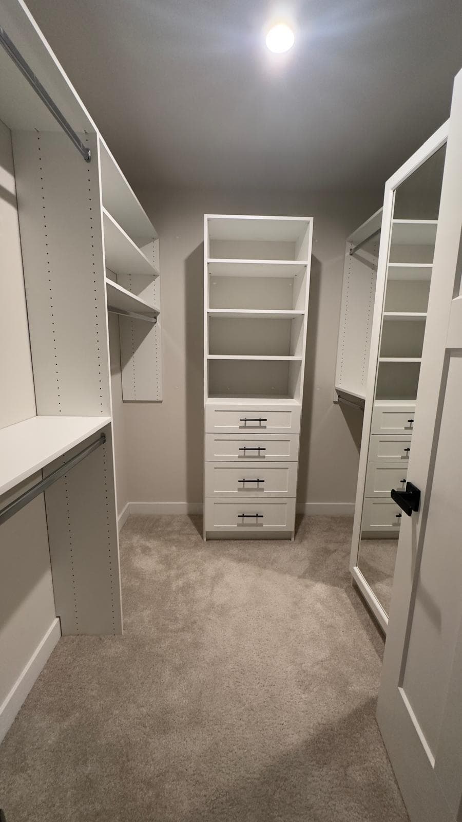 Narrow white walk-in closet with central open-shelf tower over a four-drawer base, side hanging rails, and a full-length mirrored door