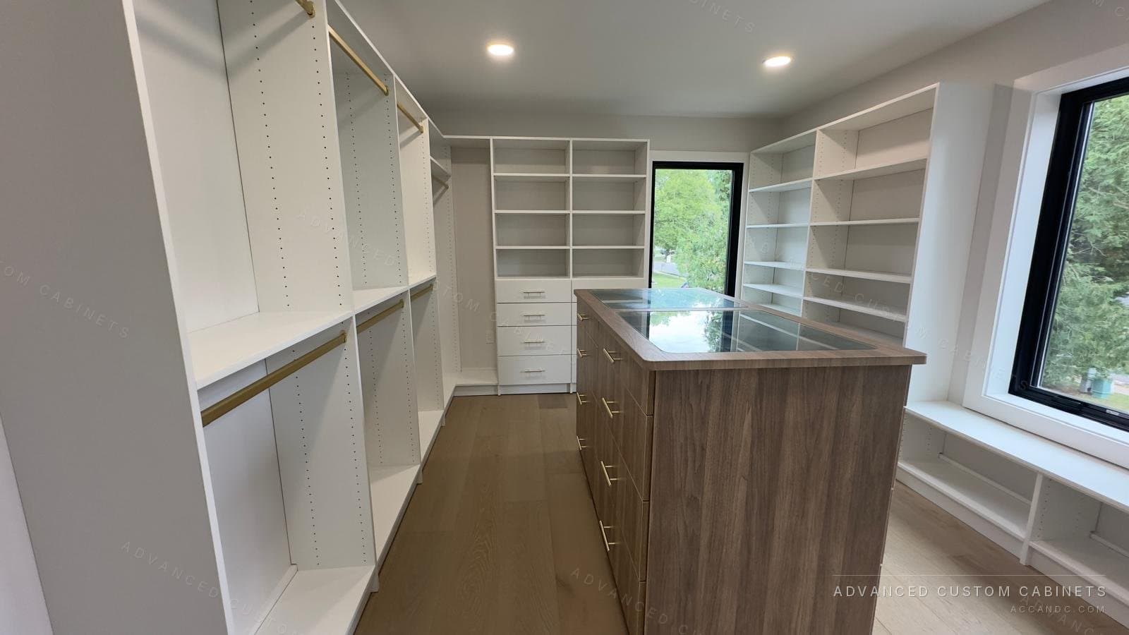 Long custom walk-in closet with a wood-front island, glass display top, brass rods, and a built-in window seat