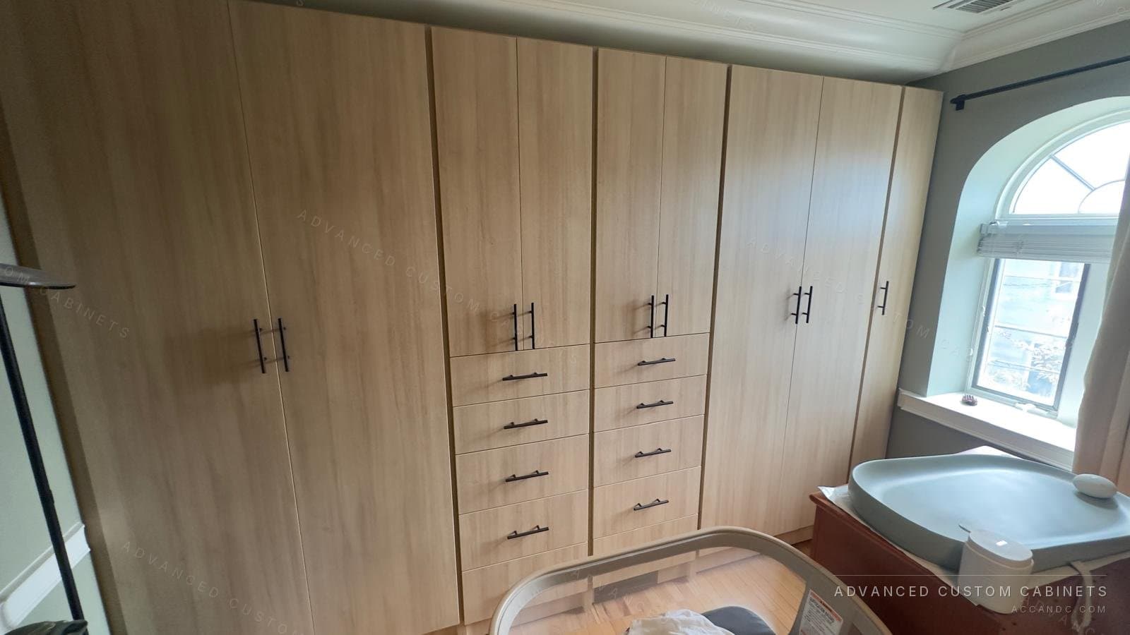 Light oak wardrobe wall with full-height doors, two drawer stacks and black bar pulls in a bedroom with an arched window