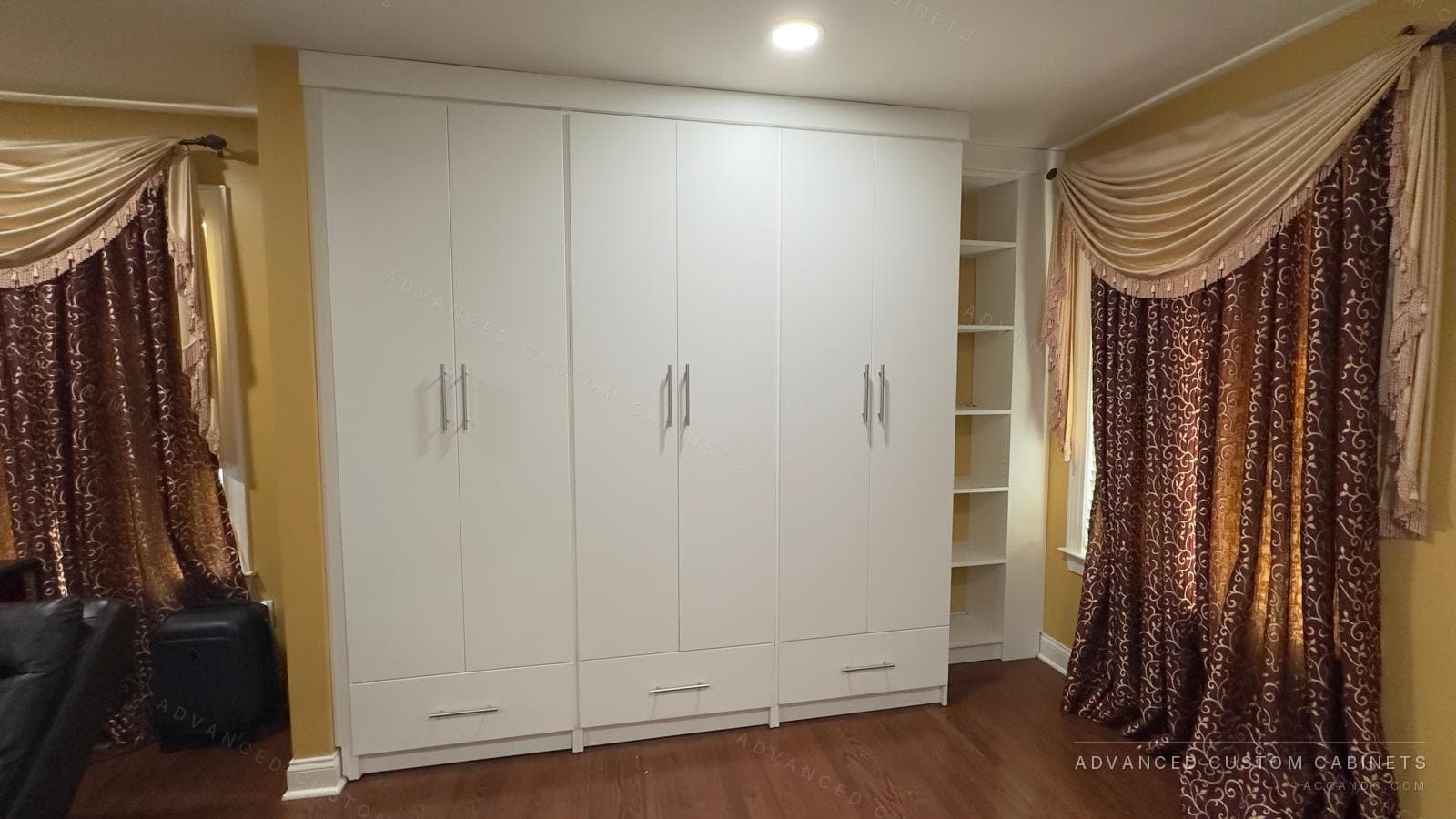 White floor-to-ceiling wardrobe wall with five doors, long bar pulls and a bottom drawer, built beside a curtained window