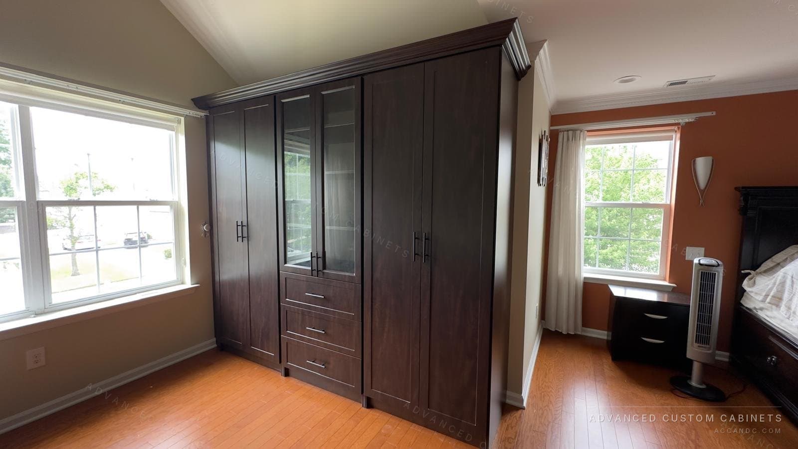 Custom espresso-finish bedroom wardrobe wall with paneled doors, a glass-front display cabinet, and a three-drawer center bank under crown molding